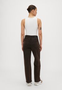 Vaqueros marrones de cintura alta con un diseño de pierna recta, tejido texturizado y dos bolsillos traseros, combinados con una blusa sin mangas blanca y zapatillas.