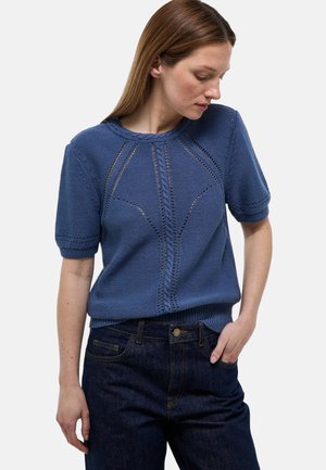 Femme portant un pull en maille bleu à manches courtes avec des trous décoratifs, associé à un jean bleu foncé, debout devant un fond uni.