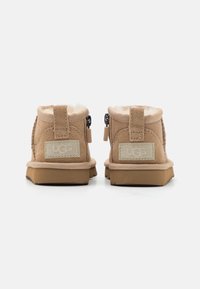 Tan suede ankle boots with shearling lining, side zipper, rubber sole, and embossed logo patch on the back. Minimalist design.