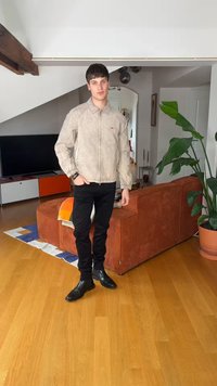 Νέος άνδρας με μπεζ μπουφάν με φερμουάρ, μαύρο παντελόνι και μαύρες μπότες στέκεται σε σαλόνι με ξύλινο πάτωμα, πορτοκαλί καναπέ, τηλεόραση και μεγάλο πράσινο φυτό.