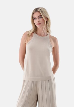 Frau mit blondem Haar, die ein ärmelloses beiges Oberteil und weite, plissierte beige Hosen trägt, steht mit den Händen hinter dem Rücken.