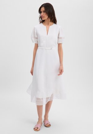 Frau trägt ein weißes Midikleid mit Puffärmeln, Spitzen-Details, Gürtel in der Taille und pinken flachen Sandalen, steht vor einem einfarbigen Hintergrund.