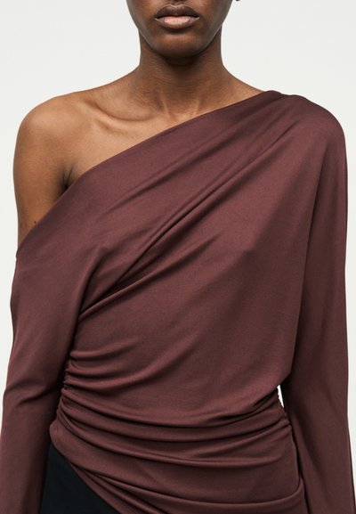 Kobieta ubrana w ciemnobrązowy, asymetryczny top z długim rękawem, z marszczonym detalem na tkaninie oraz odsłoniętym lewym ramieniem i obojczykiem.