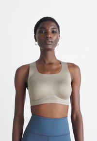Svart kvinne iført beige trenings-BH med ventilasjonshull og høyhalsede blå leggings, stående mot en enkel hvit bakgrunn.