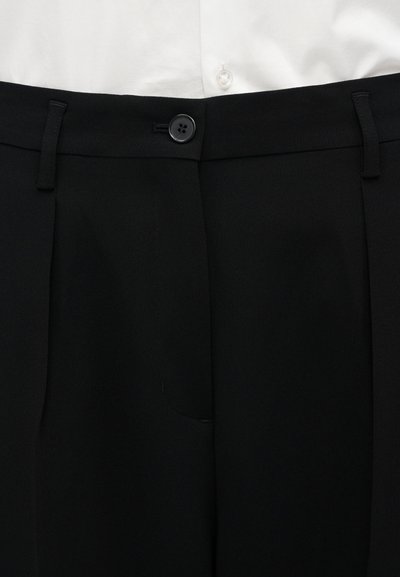 Μαύρο παντελόνι ραμμένο με θηλιές για ζώνη και ένα μαύρο κουμπί, φορεμένο με λευκό πουκάμισο με κουμπιά που είναι μπήκε μέσα.