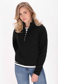 Pullover in pile nera con chiusura zip, collo alto, maniche lunghe e vestibilità slim. Indossato sopra una camicia a righe; presenta un branding minimale.