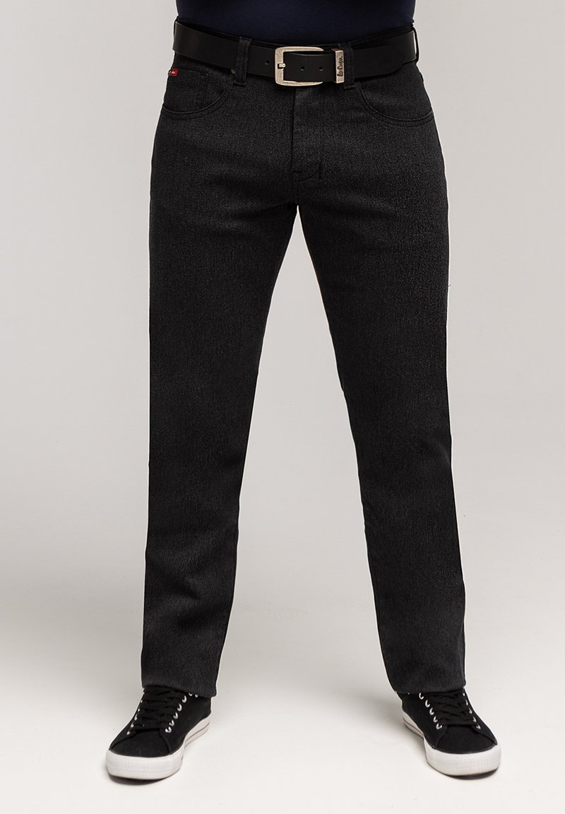 Mężczyzna ubrany w czarne dopasowane jeansy z czarnym paskiem i czarne trampki stoi na jednolitym tle.