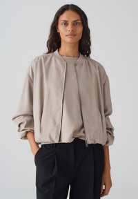 Beige bomberjack met een ritssluiting aan de voorkant, wijde mouwen en geribbelde manchetten; gecombineerd met een bijpassend coltrui en zwarte broek.