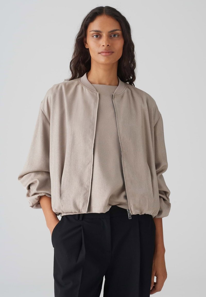 Beige bomberjack met een ritssluiting aan de voorkant, wijde mouwen en geribbelde manchetten; gecombineerd met een bijpassend coltrui en zwarte broek.