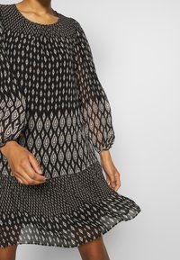 Svart klänning med rund halsringning, långa puffärmar och skiktad nederkant. Har ett diamantsmönster i kräm och dämpade nyanser genom hela plagget.