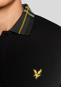 Κοντινό πλάνο ενός μαύρου πόλο με ριμπ γιακά με κίτρινες και γκρι ρίγες, και ένα μικρό κίτρινο κεντημένο λογότυπο πτηνού στο στήθος.