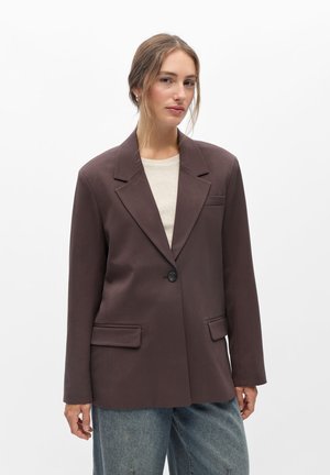 Jeune femme portant un blazer marron foncé à un seul bouton sur une chemise claire et un jean bleu, debout devant un fond blanc uni.