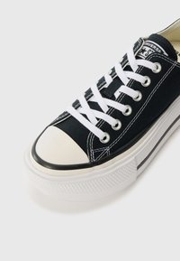 Black canvas sneaker with white laces, rubber toe cap, and textured white sole. Features a stitched design and black accent stripe.