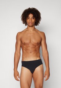 Schwarze Bikini-Höschene aus glattem Stoff, mit einem tief sitzenden Bund und minimaler Abdeckung. Das Model steht in einer neutralen Pose.