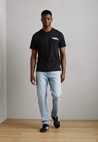 Black cotton t-shirt with white printed logo, light blue denim jeans, and black sneakers with white stripes, standing on a wooden floor.