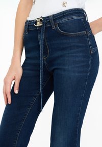 Jeans en denim bleu foncé avec une ceinture en tissu nouée à la taille, portés avec un haut blanc, et une main reposant près de la cuisse.