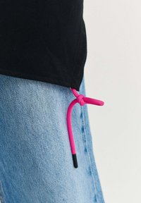 Černé tričko s růžovým tkaničkou uvázaným na lemu přes světle modré džíny na jednobarevném pozadí.