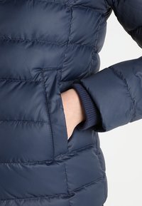 Námořnická péřová bunda s prošívaným designem, boční kapsou a žebrovanými manžetami. Materiál vypadá hladký a zateplený pro udržení tepla.