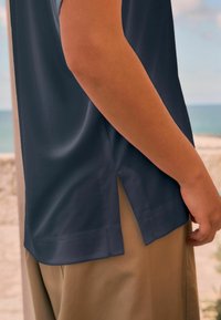 Chemise à manches courtes bleu marine avec une coupe décontractée, présente des fentes latérales à l'ourlet et une texture lisse, assortie à un pantalon léger en kaki.
