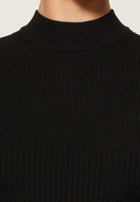 Gros plan d'un pull noir à col roulé côtelé porté sur le cou et le haut de la poitrine d'une personne, montrant la texture du tissu et la forme du col.