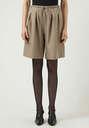Shorts beige a vita alta con pieghe e coulisse, indossati con calze nere trasparenti e tacchi neri con punta a mandorla su una persona in piedi.