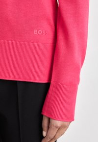 Svetlo roza pulover z rebrastimi manšeti in robom. Majhna, vgrajena oznaka "BOSS" v pletenini. Gladka tekstura in prilegajoča se zasnova.