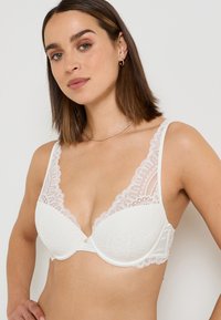 Soutien-gorge en dentelle blanche avec un décolleté plongeant, des bords festonnés et des bonnets rembourrés. Présente un petit détail décoratif au centre.