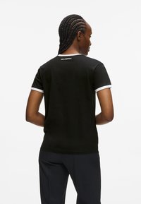 Czarny T-shirt z białym kołnierzykiem i akcentami na rękawach, z logo z tyłu. Materiał wydaje się gładki i dopasowany.