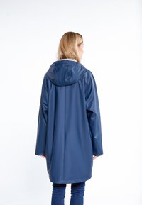 Szőke hajú nő, aki sötétkék kapucnis esőkabátot és sötét farmernadrágot visel, háttal álló helyzetben egy fehér háttér előtt.