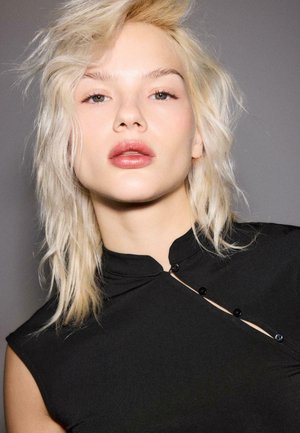 Giovane con capelli biondo chiaro e espressione neutra, che indossa una canottiera nera con dettagli di bottoni diagonali su uno sfondo grigio.