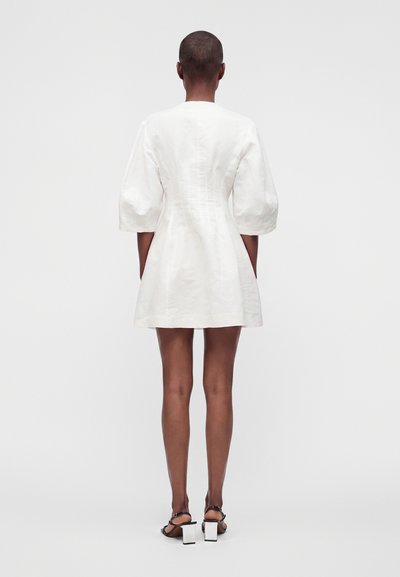 Personne portant une robe courte blanche à manches bouffantes et des sandales noires à talons blocs blancs, debout de dos sur un fond uni.