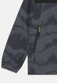 Giacca grigia con un motivo testurizzato, caratterizzata da un pannello nero e una tasca con zip con un tirante giallo. Polsini elastici sulle maniche.