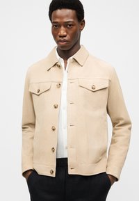 Νεαρός άνδρας που φοράει μπεζ σακάκι με κουμπιά πάνω από άσπρο πουκάμισο, με τα χέρια στις τσέπες μαύρου παντελονιού, μπροστά σε απλό φόντο.