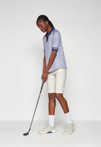 Short-sleeve golf polo, blue with pattern, paired with light beige shorts. Gray athletic shoes with mint accents. Golf club in hand.