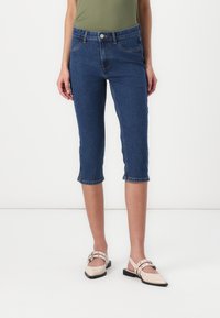 Pantalons capri en denim bleu foncé, avec une taille ajustée, un design à cinq poches et des revers roulés, associés à des chaussures beiges à enfiler.