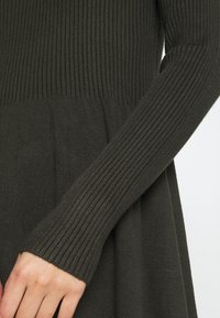 Robe tricotée vert olive foncé avec une texture côtelée, dotée de manches longues et d'un bodice ajusté qui s'évasent vers l'ourlet.