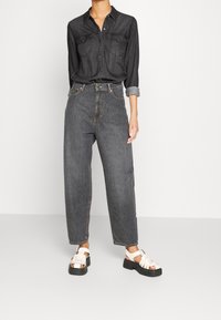 Camicia in denim nera con bottoni frontali e due tasche sul petto, abbinata a jeans grigi scuri a vita alta e dalla vestibilità ampia, e sandali bianchi.