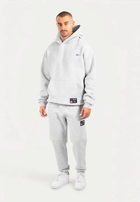 Grijze hoodie en joggingbroek set gemaakt van zachte stof. Heeft een logo op de borst, zijzakken en een patch-detail op de joggingbroek. Witte sneakers gedragen.