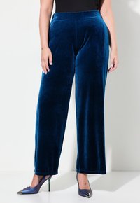 Sammetsoveralls med vida ben i djupblått, med en mjuk textur och hög midja, parat med klackskor med spetsig tå och metalliska detaljer.