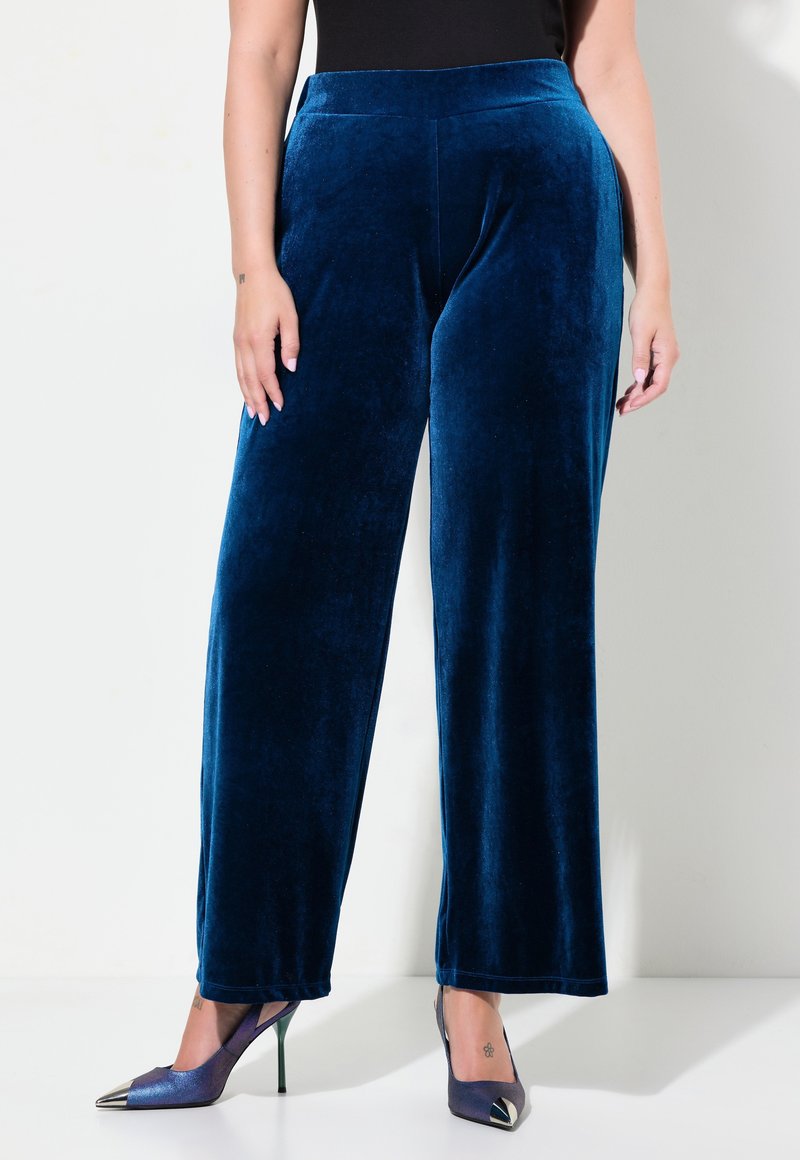 Sammetsoveralls med vida ben i djupblått, med en mjuk textur och hög midja, parat med klackskor med spetsig tå och metalliska detaljer.
