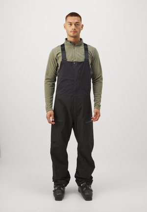 Pantalones de esquí tipo peto en tela negra con tirantes ajustables y bolsillos con cremallera en el pecho. Llevados sobre una camiseta de manga larga verde, combinados con botas negras.