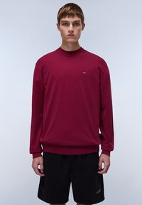 Suéter de punto burdeos con mangas largas y cuello acanalado, que presenta un pequeño logo en el pecho y un corte relajado. Combinado con pantalones cortos oscuros.