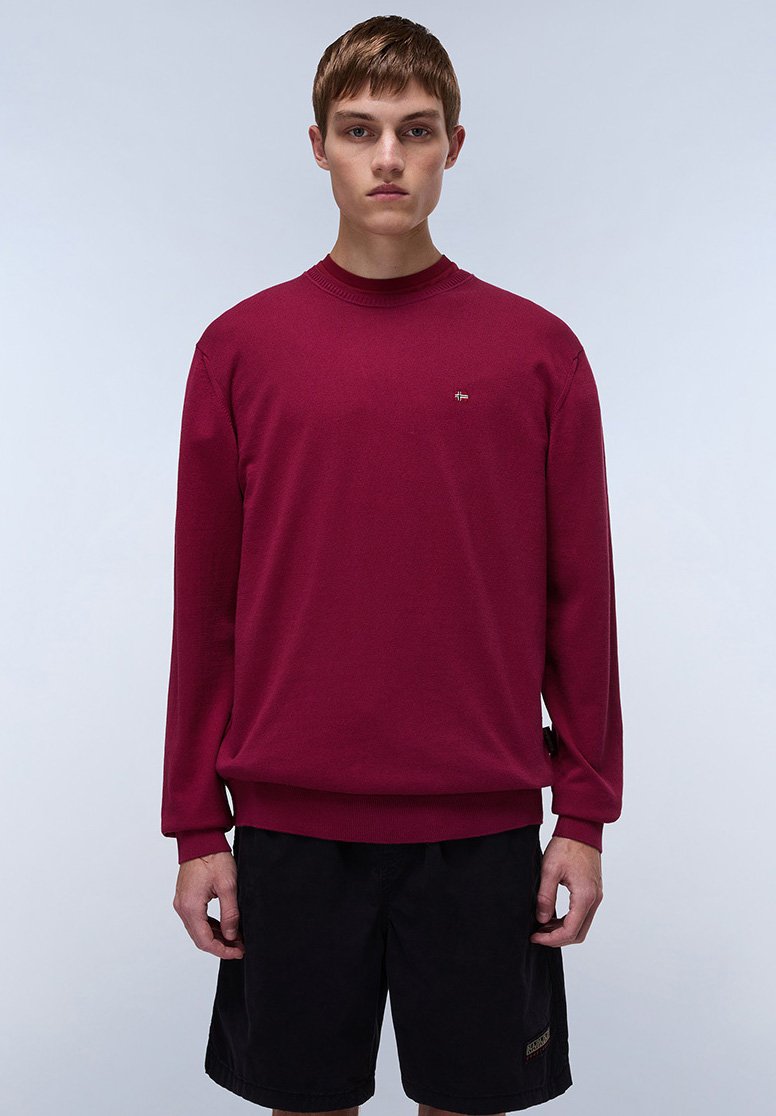 Suéter de punto burdeos con mangas largas y cuello acanalado, que presenta un pequeño logo en el pecho y un corte relajado. Combinado con pantalones cortos oscuros.