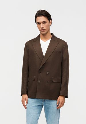 Mladý muž v tmavohnedom dvojradovom saku, bielej košeli a svetlomodrých džínsoch stojaci pred jednoduchým svetlým pozadím.