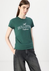 Zelené bavlněné tričko s kulatým výstřihem, na kterém je bílý nápis "HOLLISTER California" a logo. V kombinaci s tmavě šedými širokými kalhotami.