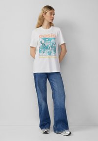 T-shirt en coton blanc avec un graphique illustré bleu et le texte orange "oasis" et "Definitely Maybe". Assorti avec un jean bleu à jambes larges.
