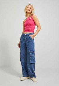 Top sin mangas asimétrico rosa ajustado combinado con pantalones cargo de denim azul holgados que cuentan con múltiples bolsillos y un ajuste relajado. Zapatillas neutras.