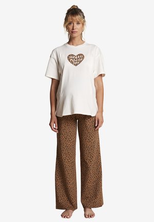 Woman standing barefoot wearing a white shirt with a leopard heart and text "Heart Full of Joy" and brown leopard print wide pants.
