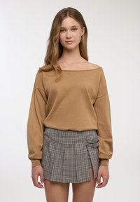 Maglione marrone lavorato a maglia con scollo a barca, abbinato a una gonna plissettata a quadri blu e beige con due fibbie laterali.