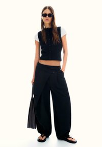 Zwart gekropt vest over een wit T-shirt, gecombineerd met wijde zwarte broek met een unieke drape. Platte zwarte sandalen maken de look compleet.